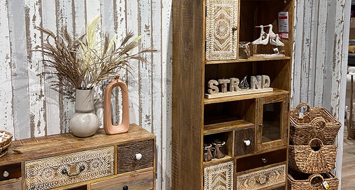 Man sieht den Ausschnitt eines Sideboards und rechts daneben eines Regals. Die Möbel sind aus rustikal aussehendem Holz gefertigt. Die Schubladen der beiden Stücke haben unterschiedliche Größen und Fronten in verschiedneen Farben. Die griffe sind ebenso in unterschiedlichen Farben gehalten. Manche der Schubladen haben ein weißes, verschnörkeltes Muster. Auf dem Sideboard stehen zwei dekorative Vasen, die linke von ihnen ist befüllt mit einem Trockenblumenstrauß. Ganz rechts im Bild stehen mehrere geflochtene Körbe, die dekorativ aufeinandergestapelt sind.
