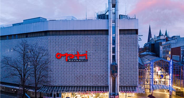 Man sieht die Fassade des neuen Opti-Wohnwelt-Cityhauses in der Innenstadt von Bremen. Auf dem Bild dämmert es, die Fenster im Erdgeschoss sind beleuchtet.