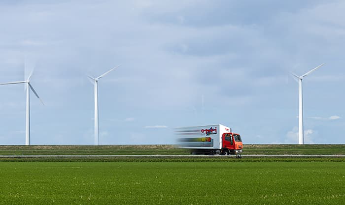 Man sieht eine Landstraße. Im Hintergrund drehen sich drei Windräder. Auf der Straße fährt ein Opti-Wohnwelt-Transporter. 