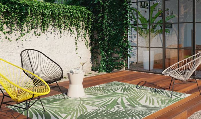 Wohnmagazin: Außenbereich mit Holzboden, darauf Outdoor-Teppich mit grünem Palmen-Muster und zwei sommerliche Flechtstühle in gelb und schwarz.