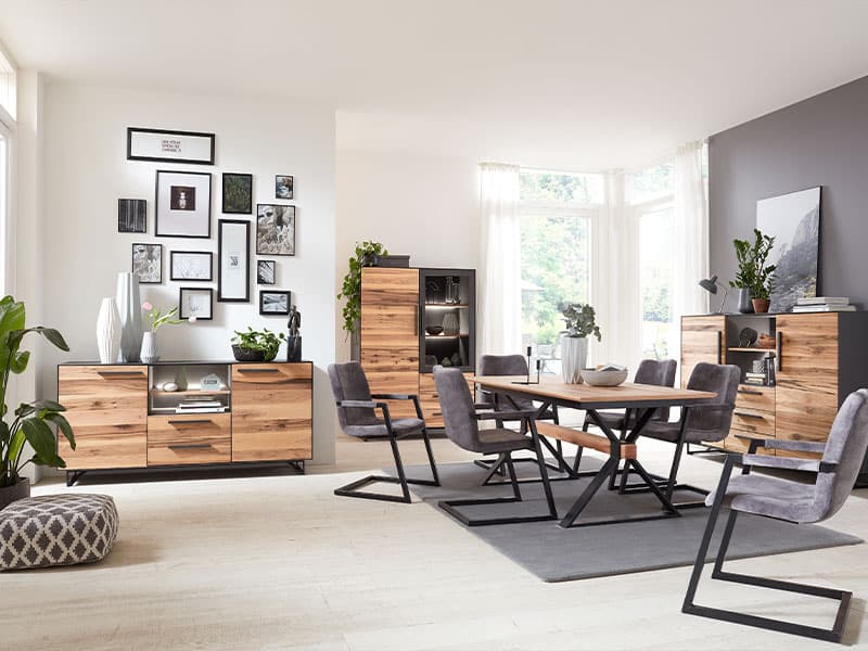 Mehrere graue Schwingstühle stehen um einem Holztisch zwischen drei Holzkommoden