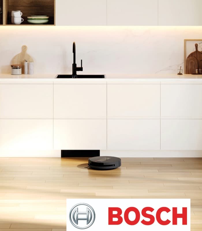Eine helle Küche mit schwarzem Waschbecken und Kochfeld. Weiterhin ist ein schwarzer Saug- & Wischroboter zu sehen. Unten rechts befindet sich ein rotes Bosch-Logo.