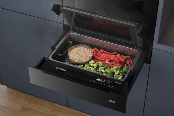 Geöffnete schwarze Küchenschublade mit Glasdeckel, gefüllt mit Gemüse, Paprika und einer Schale Getreide.