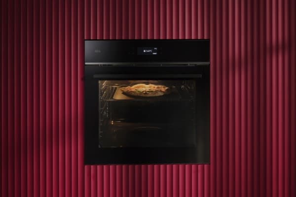 Schwarzer Einbaubackofen mit einer Pizza im Garraum vor einer bordeauxroten geriffelten Wand.