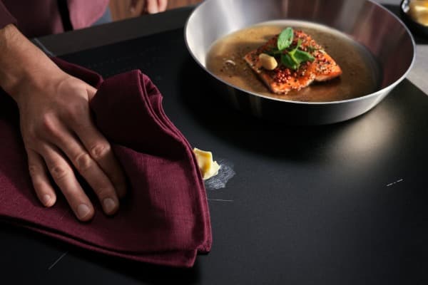 Hand wischt mit einem bordeauxroten Tuch eine verschmutzte schwarze Kochfeldoberfläche neben einer Pfanne mit gebratenem Lachs ab.