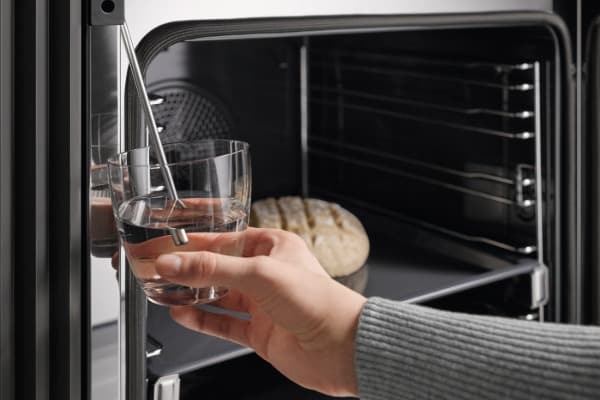Hand füllt ein Glas am Wasserauslauf eines geöffneten Dampfbackofens; im Garraum liegt ein Brot.