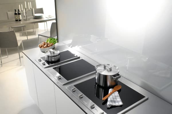 Helle, moderne Küche mit mehreren schwarzen Induktionskochfeldern und Edelstahltöpfen auf einer weißen Küchenzeile.