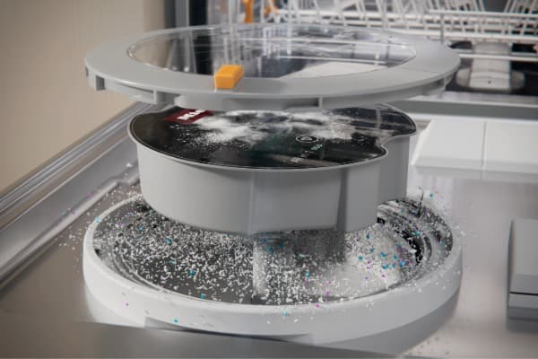 Nahaufnahme eines runden Bauteils mit transparentem Deckel im Inneren einer Geschirrspülmaschine, umgeben von Wasser und Spülmittel.