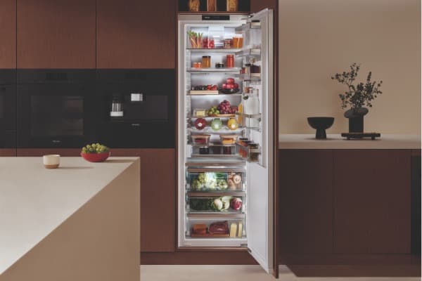 Geöffneter, voll eingeräumter Kühlschrank mit Obst, Gemüse, Getränken und Lebensmitteln in einer modernen Küche mit dunklen Holzfronten.
