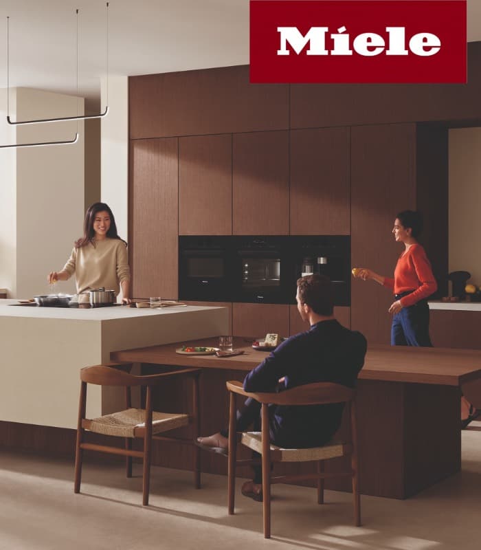 Moderne Miele-Küche mit dunklen Holzfronten und Kücheninsel; eine Frau kocht, eine weitere Frau steht daneben und ein Mann sitzt am Esstisch.